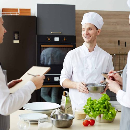 Cheerful Optimistic Handsome Chef In Hat Holding Bowl With Whisk While Sharing Cooking Ideas While Preparing New Recipe Together With Colleagues At Commercial Kitchen